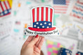 Load image into Gallery viewer, Hand holding a 'Happy Constitution Day' sticker with an American flag design against a blurred background of flags.