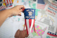 Load image into Gallery viewer, Hand holding a DIY windsock with 'We the People' design against a colorful background