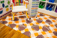 Load image into Gallery viewer, A children's play rug with an orange and white checkered pattern and cartoon acorns wearing plaid scarves, shown in a playroom setting with a white high chair and storage shelves.
