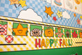 Load image into Gallery viewer, A close-up of a colorful autumn-themed wall mural or classroom display with the words “HAPPY FALL!” in large multicolored letters. The design includes cartoon maple leaves, pumpkins, a scarecrow, clouds, stars, and a blue-and-white checkered background.