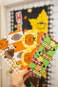 Load image into Gallery viewer, A hand with red nail polish holds four colorful autumn-themed paper envelopes or cards in front of a blurred fall classroom decoration background. The designs feature pumpkins, acorns, leaves, and checkered patterns in orange, green, brown, yellow, and white.