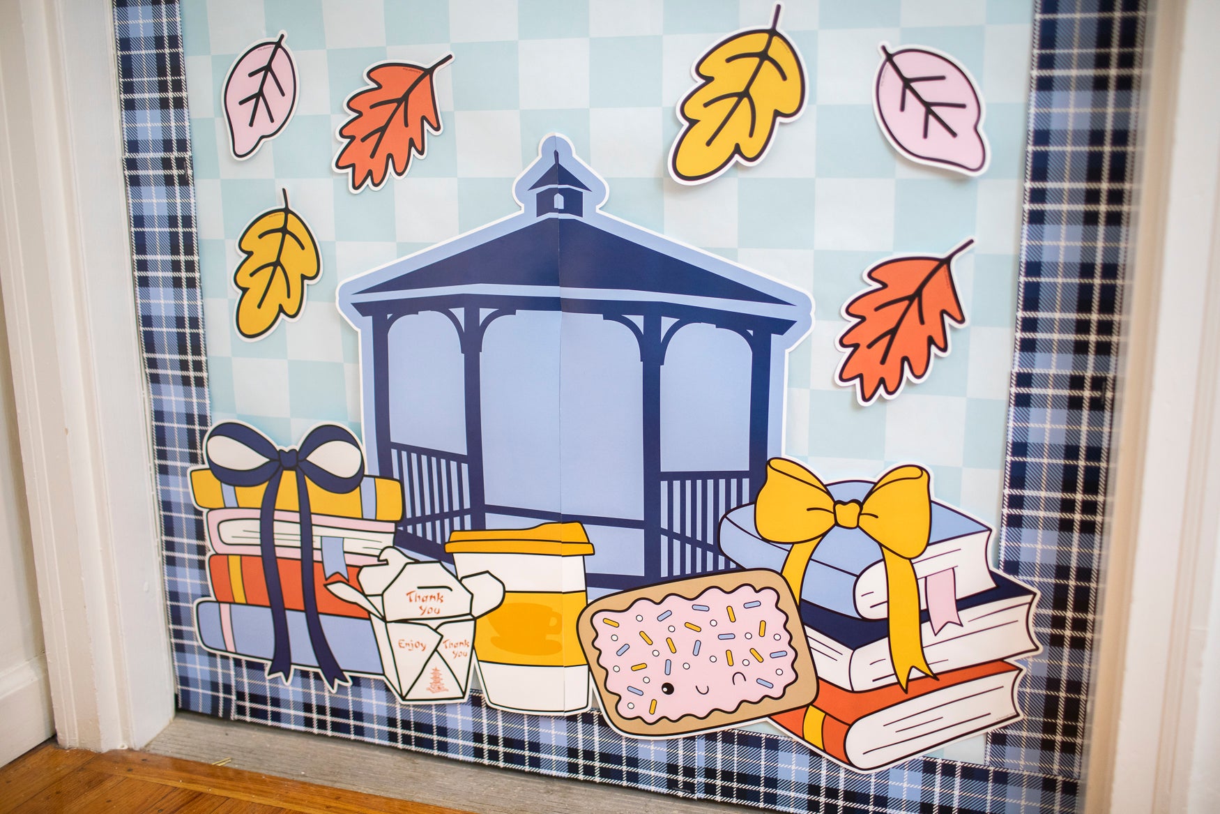 A decorative autumn-themed window decal or sticker sheet featuring a blue gazebo, gift boxes with bows, books, a coffee cup, a pastry, and scattered fall leaves on a light blue check background.