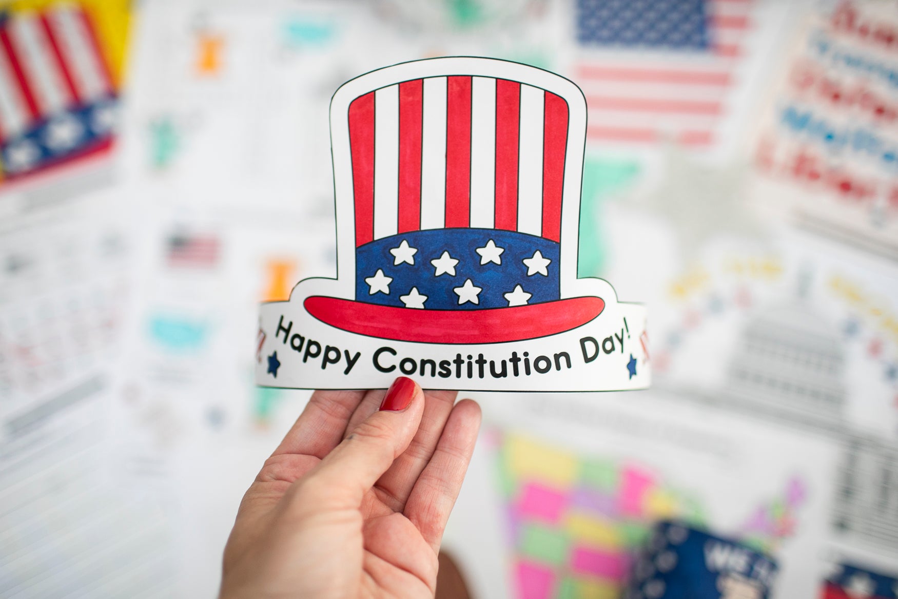 Hand holding a 'Happy Constitution Day' sticker with an American flag design against a blurred background of flags.