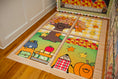 Load image into Gallery viewer, A wooden children's rug with a four-panel autumn-themed design, placed on a hardwood floor next to a white paneled wall. The rug features cartoon illustrations of a tree with colorful leaves, a pumpkin, a checkered character, and a stack of books with stars.
