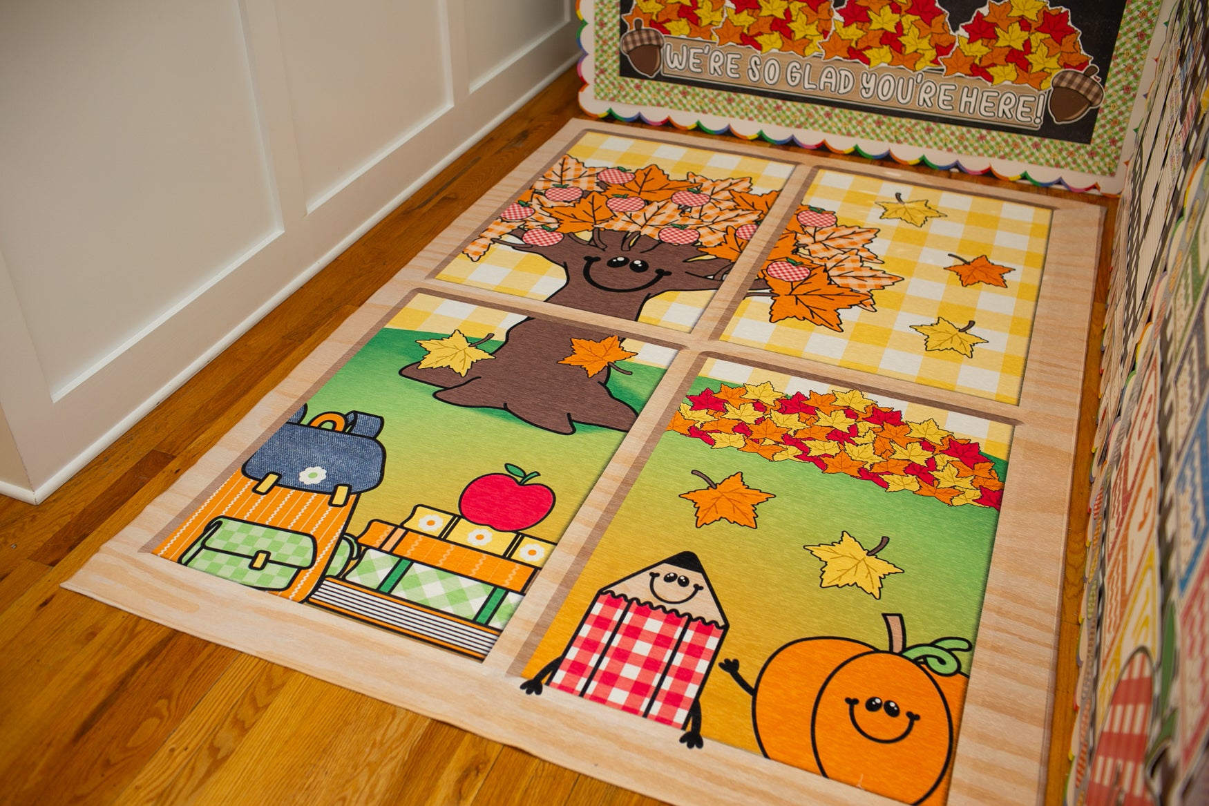A wooden children's rug with a four-panel autumn-themed design, placed on a hardwood floor next to a white paneled wall. The rug features cartoon illustrations of a tree with colorful leaves, a pumpkin, a checkered character, and a stack of books with stars.