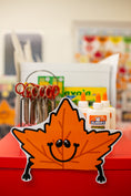 Load image into Gallery viewer, A smiling orange autumn leaf sign with black eyes and arms stands on a red surface in front of a craft station setup. A white glue bottle labeled Elmer's is visible behind it, along with scissors in a holder and a blurred indoor background.
