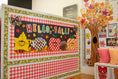 Load image into Gallery viewer, A decorative autumn-themed classroom display board with the words 'HELLO, FALL!' in colorful letters, featuring smiling fall icons like a yellow leaf, a plaid pumpkin, an apple, and an acorn. The board is framed in red-and-white gingham and patterned borders, shown in a school-like interior with a tree cutout and colorful pillows nearby.
