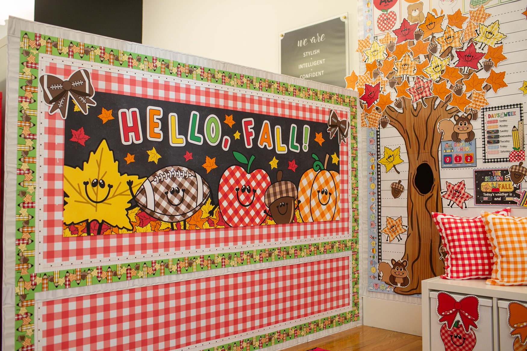 A decorative autumn-themed classroom display board with the words 'HELLO, FALL!' in colorful letters, featuring smiling fall icons like a yellow leaf, a plaid pumpkin, an apple, and an acorn. The board is framed in red-and-white gingham and patterned borders, shown in a school-like interior with a tree cutout and colorful pillows nearby.