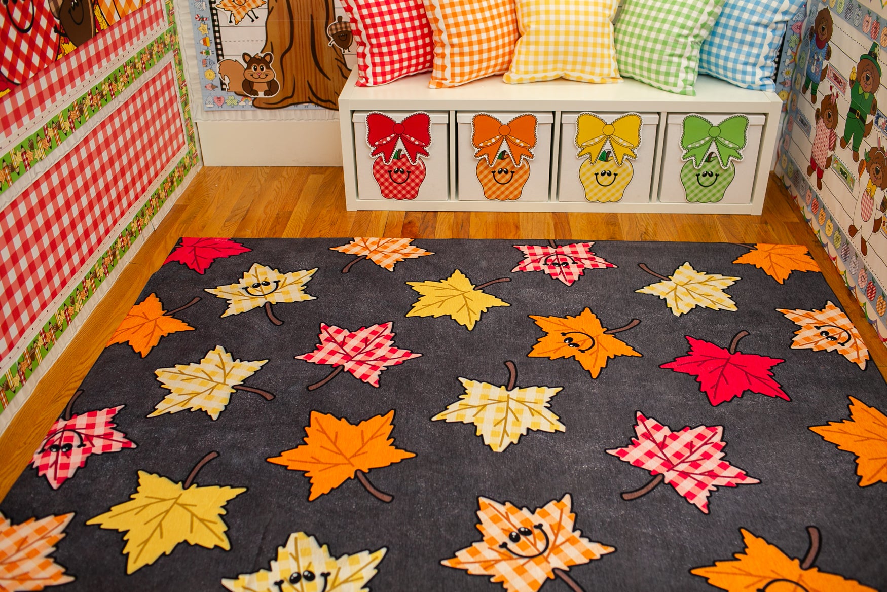 A dark gray area rug with a playful pattern of colorful autumn maple leaves in red, orange, yellow, and white, styled in a children's room with a matching white storage bench and gingham pillows.