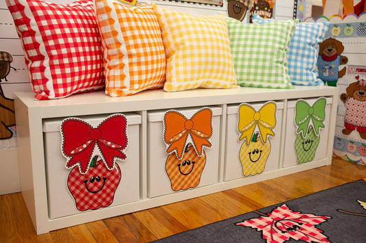 A white storage bench with four drawers decorated with colorful apple faces and bows, topped with six gingham throw pillows in red, orange, yellow, green, and blue, set in a child's room.