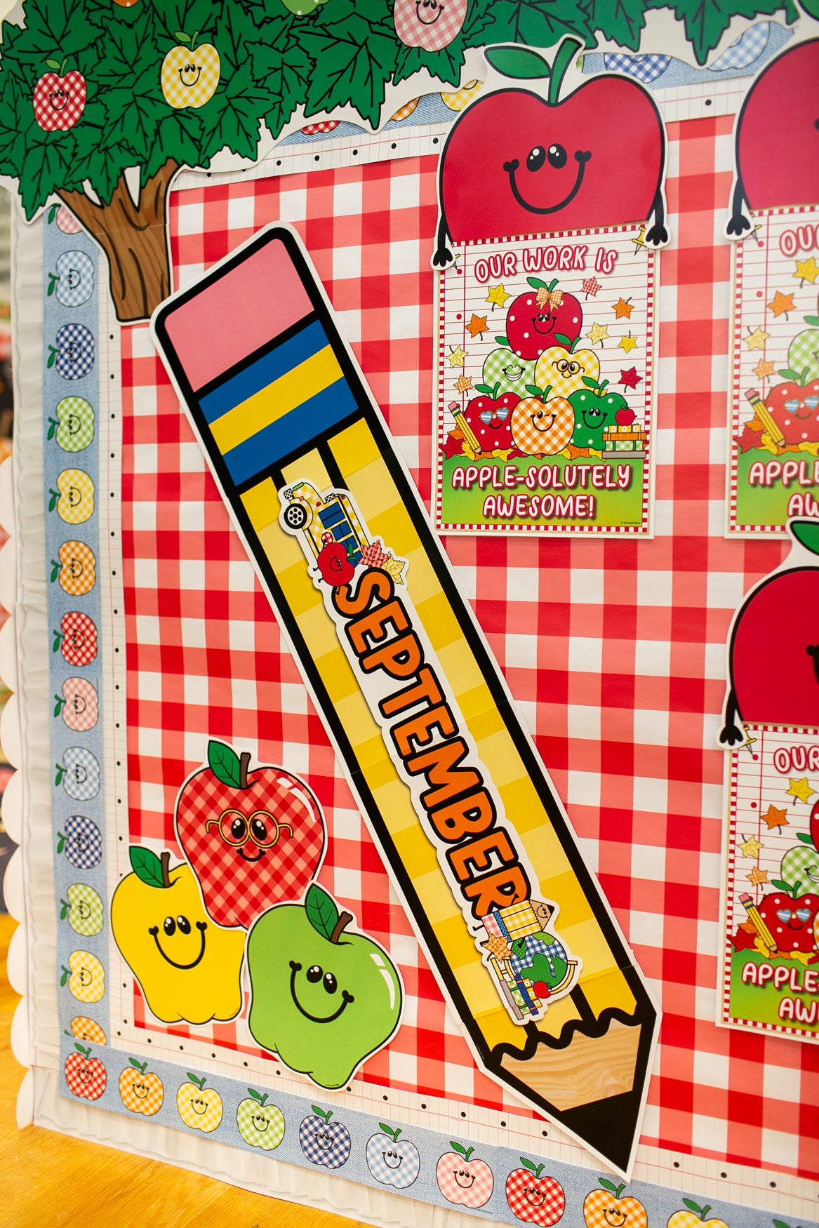 A close-up of a colorful school-themed bulletin board or classroom display featuring a large yellow pencil with 'SEPTEMBER' written on it, red apple graphics, smiling apples, and a red-and-white gingham background.