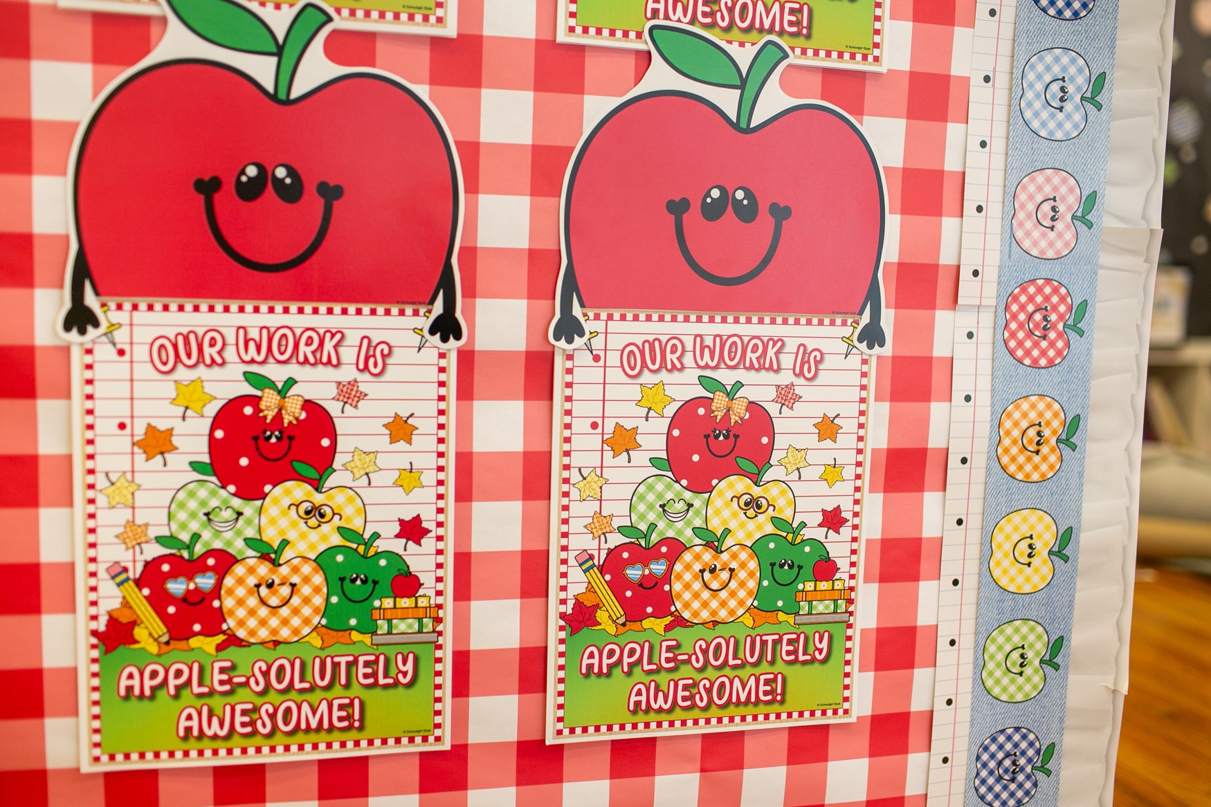 A close-up of a decorative educational poster or chart featuring two large red smiling apples above colorful cartoon apples and the phrase 'Our Work Is Apple-Solutely Awesome!' on a red-and-white checkered background.