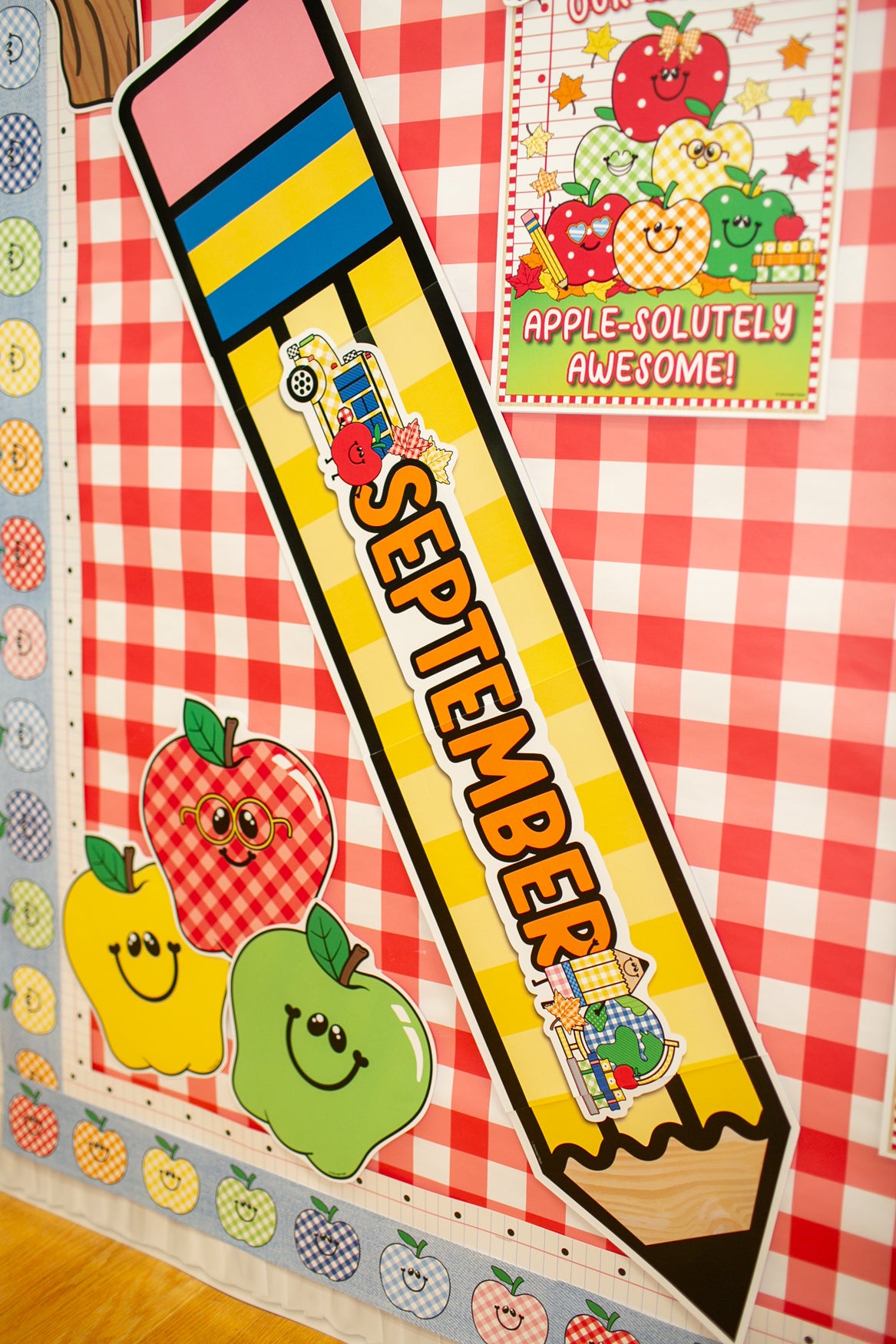 A large decorative pencil-shaped sign reading “SEPTEMBER” is displayed on a red-and-white checkered classroom-style wall with apple illustrations and a small sign that says “APPLE-SOLITELY AWESOME!”.
