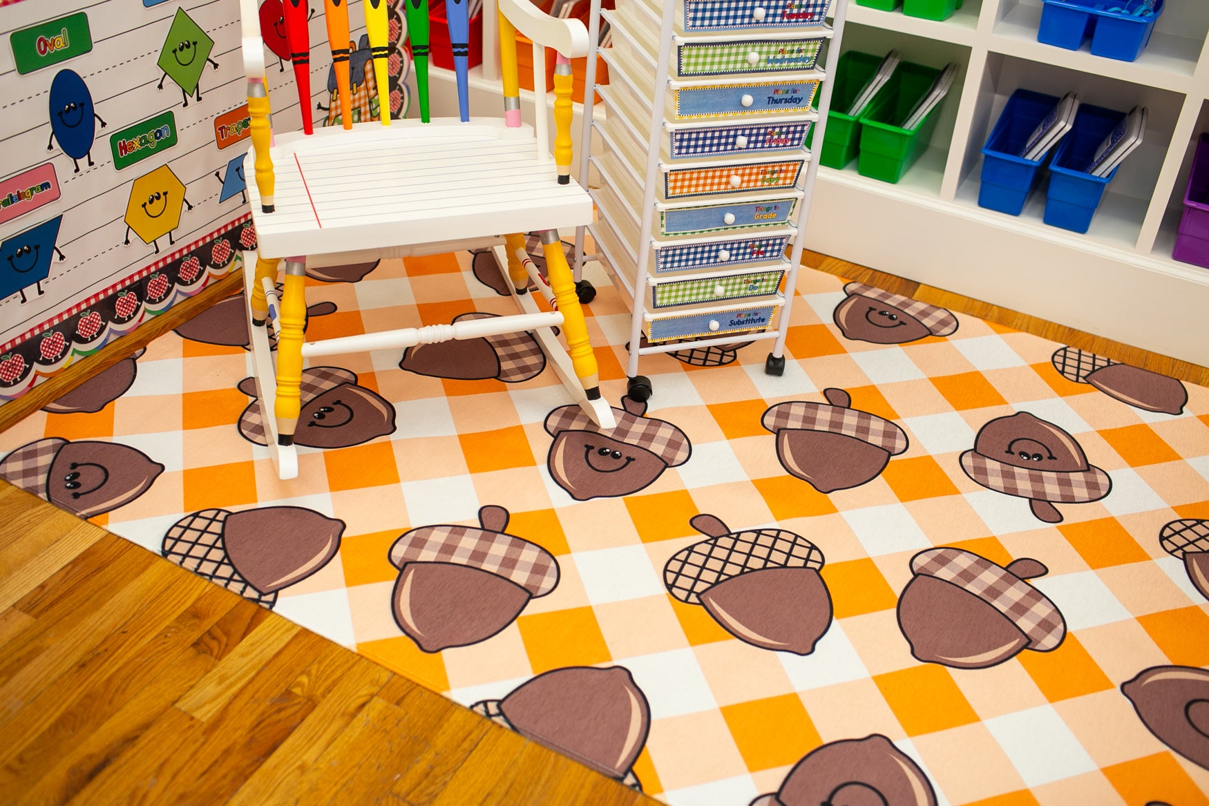A children's play rug with an orange and white checkered pattern and cartoon acorns wearing plaid scarves, shown in a playroom setting with a white high chair and storage shelves.