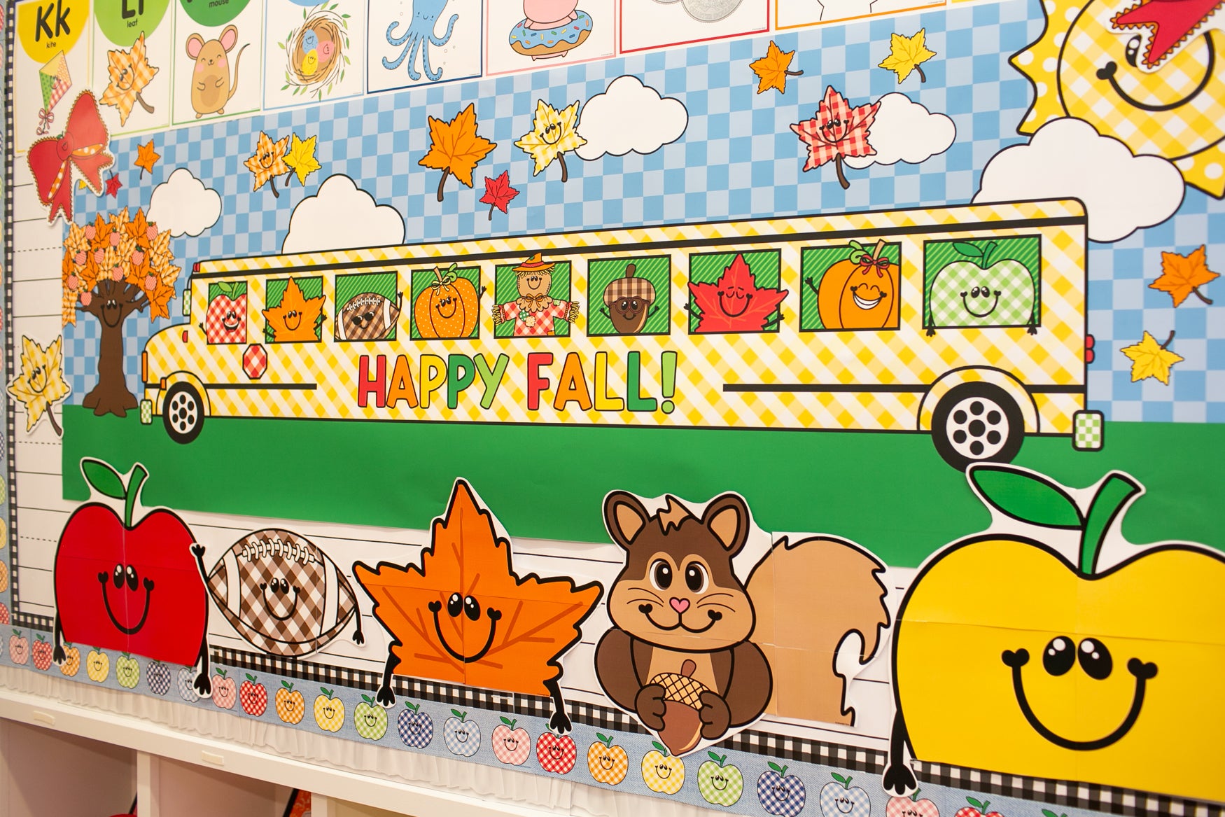 A colorful autumn-themed classroom wall display featuring a yellow school bus with 'HAPPY FALL!' text, smiling apples, a leaf, a squirrel, clouds, stars, and a blue-and-white checkered sky.