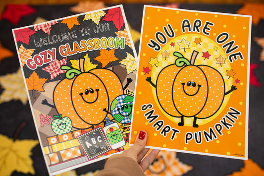 Two autumn-themed printable classroom posters held up by a hand. The left poster reads 'Welcome to our Cozy Classroom' with a smiling orange pumpkin, fall leaves, and school icons.