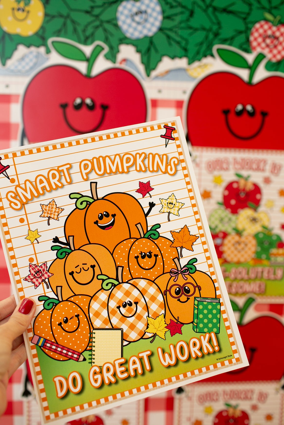 A hand holds a colorful autumn-themed notebook or planner cover featuring smiling pumpkins, stars, and the text 'SMART PUMPKIN DO GREAT WORK!'. The background shows more matching red apple and fall-themed stationery items.