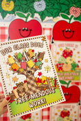 Load image into Gallery viewer, A hand with red nail polish holds a printed card or notebook cover featuring a cartoon tree with colorful leaves and the text 'OUR CLASS DOES TREE-MENDOUS WORK!'. The background shows more similar apple-themed printed materials.
