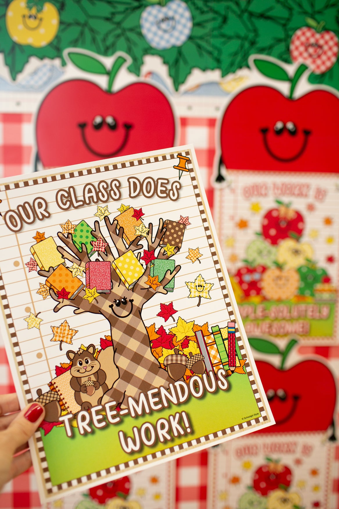 A hand with red nail polish holds a printed card or notebook cover featuring a cartoon tree with colorful leaves and the text 'OUR CLASS DOES TREE-MENDOUS WORK!'. The background shows more similar apple-themed printed materials.