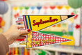 Load image into Gallery viewer, A hand with red nail polish holds two decorative pennant flags shaped like pencils. The top flag is yellow with the word 'September' in red script, and the bottom flag is red-and-white gingham with 'Hello September' in yellow.
