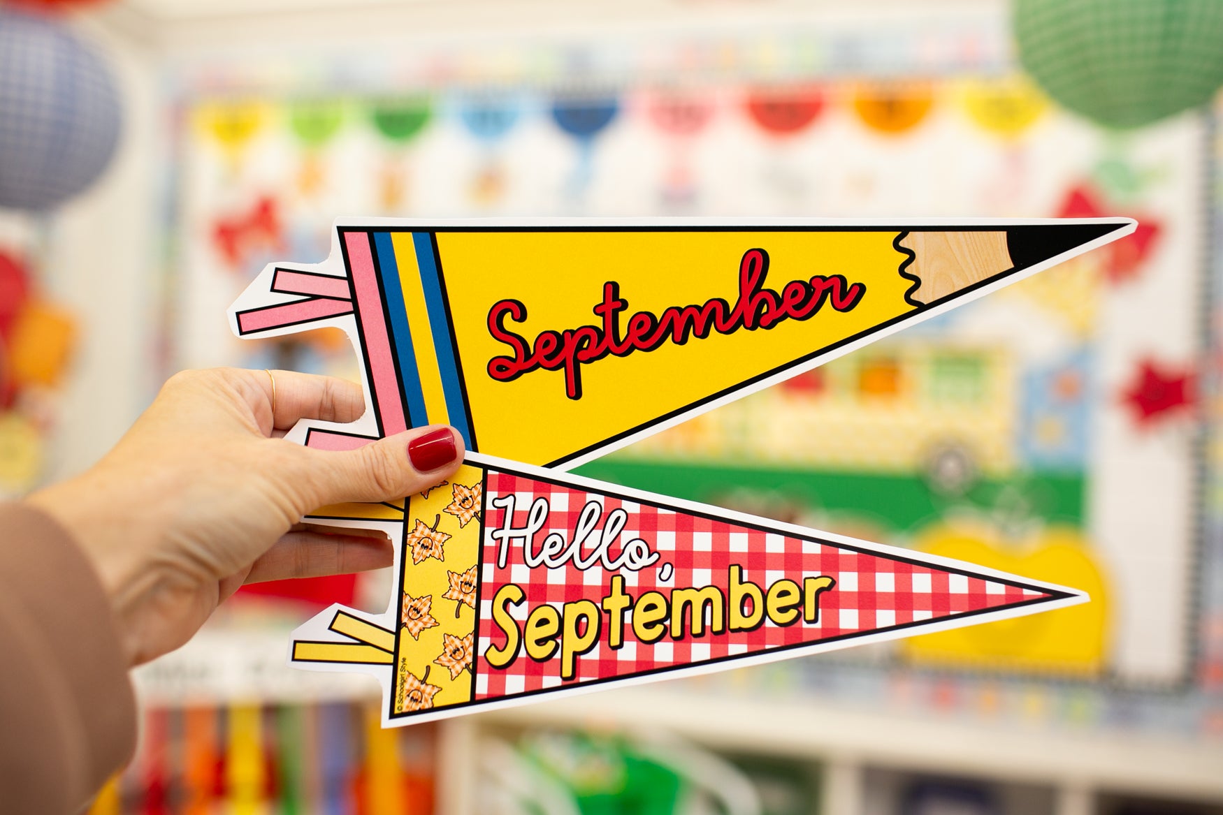 A hand with red nail polish holds two decorative pennant flags shaped like pencils. The top flag is yellow with the word 'September' in red script, and the bottom flag is red-and-white gingham with 'Hello September' in yellow.