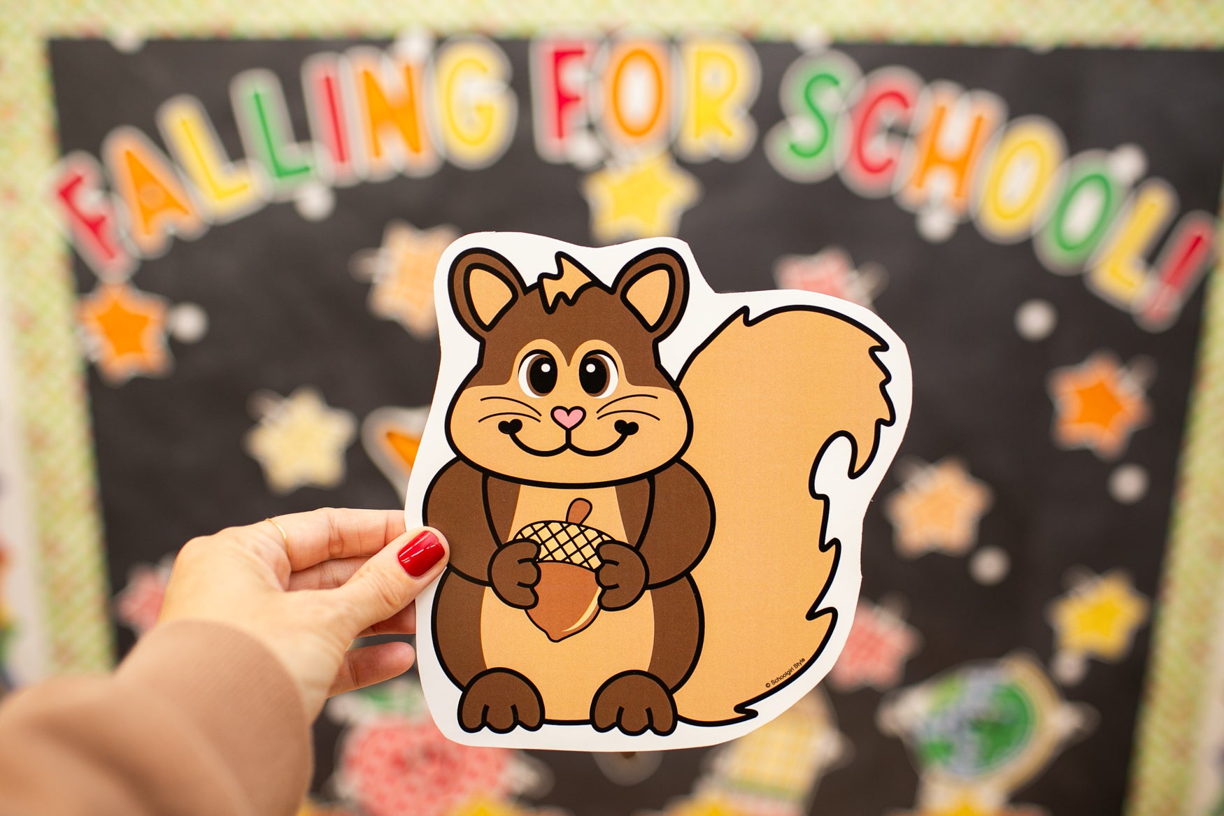 A hand with red nail polish holds a die-cut sticker of a cartoon squirrel holding an acorn in front of a school-themed 'FALLING FOR SCHOOL!' bulletin board.