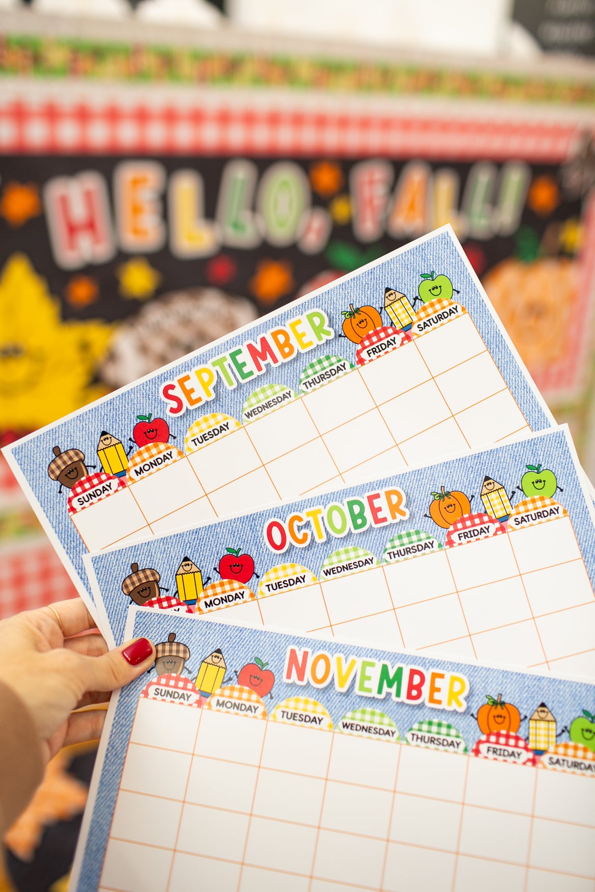 Three colorful monthly planner inserts for September, October, and November are held by a hand with red nail polish. The inserts feature a blue grid layout with cute autumn-themed illustrations of pumpkins, apples, and fall foods along the top border, set against a blurred 'HELLO, FALL!' classroom background.