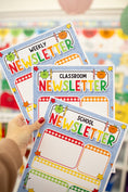 Load image into Gallery viewer, A hand with red nail polish holds three colorful printed newsletters labeled Weekly, Classroom, and School Newsletter. The newsletters feature playful autumn-themed graphics including pumpkins, stars, and checkered borders, set against a softly blurred classroom background.
