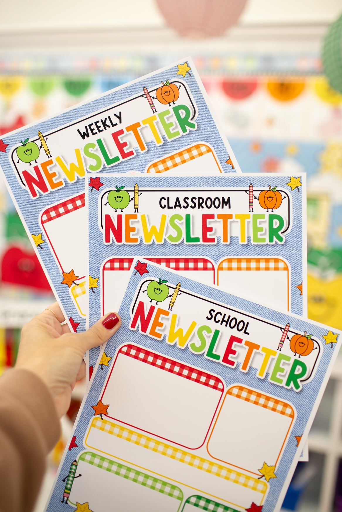 A hand with red nail polish holds three colorful printed newsletters labeled Weekly, Classroom, and School Newsletter. The newsletters feature playful autumn-themed graphics including pumpkins, stars, and checkered borders, set against a softly blurred classroom background.
