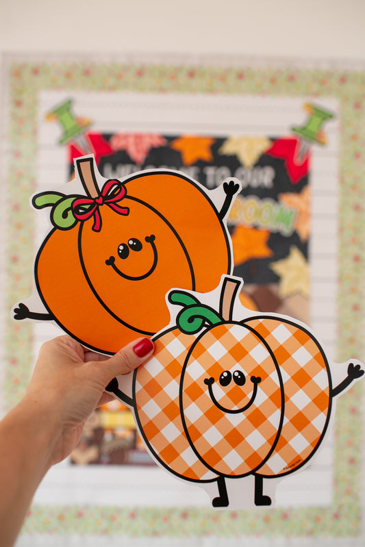A hand with red nail polish holds two die-cut pumpkin stickers in front of a blurred autumn-themed print. The stickers are orange with black outlines, smiling faces, and one has a green leaf stem with a pink bow, while the other has an orange and white gingham pattern with a green leaf stem.