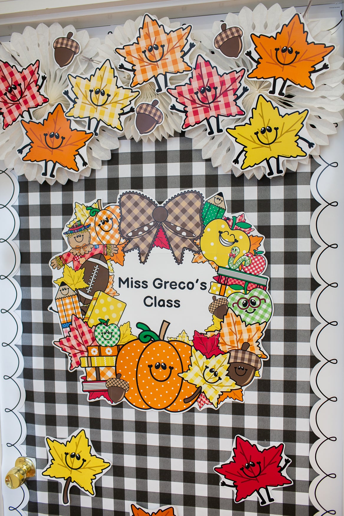 A fall-themed classroom bulletin board display featuring a black and white gingham background, a central wreath with pumpkins, acorns, bows, and autumn decorations, and the text 'Miss Greco's Class.' Decorative paper maple leaves and acorns hang around the top and bottom. Editable printable classroom decor wreath. 